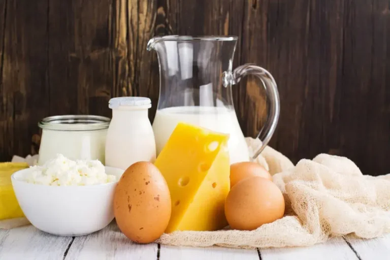 A variety of dairy products including a jug of milk, a bottle of milk, a bowl of cottage cheese, a block of Swiss cheese, and brown eggs on a white tablecloth with a rustic wooden background.