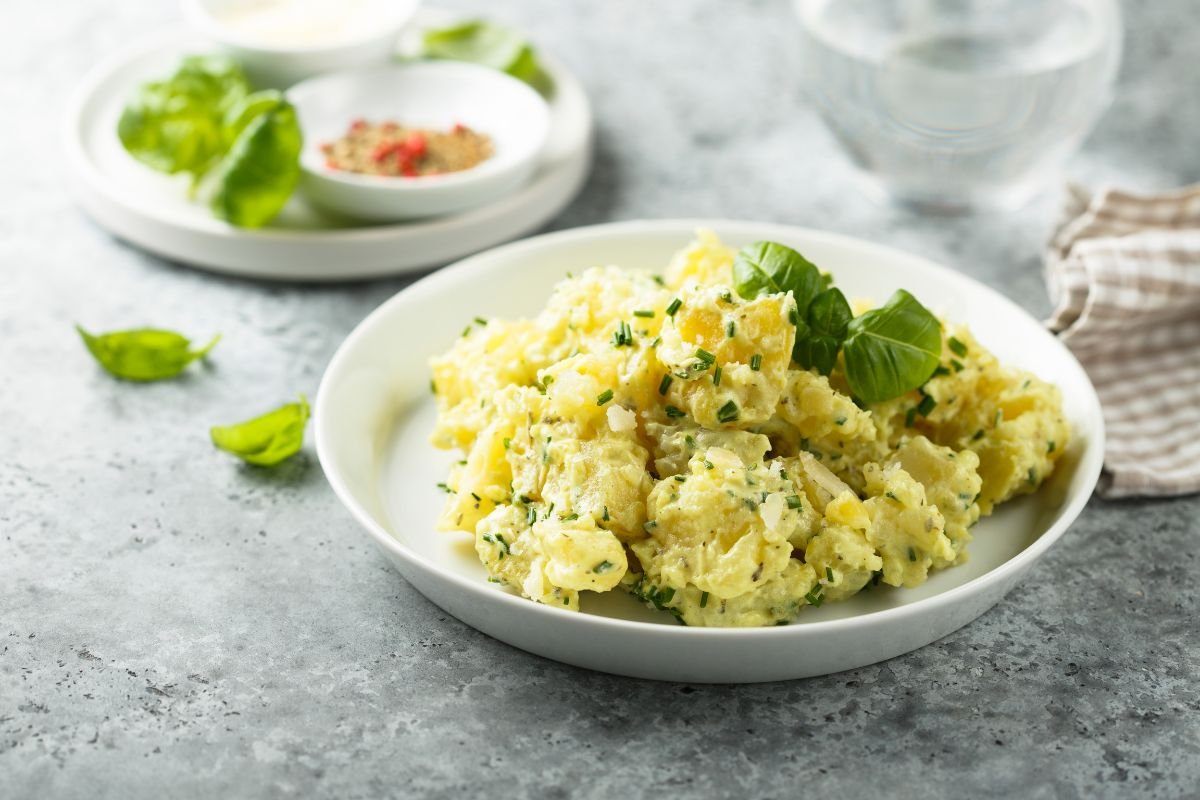 A classic Southern-style potato salad garnished with fresh green herbs, served in a white bowl against a gray textured background with basil leaves, a water glass, and a folded cloth napkin in the backdrop.